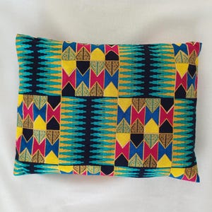Peut inclure: Coussin rectangulaire avec un imprimé africain vibrant. Le coussin présente un motif répétitif de formes géométriques turquoise, jaunes, roses et noires. Le coussin est posé sur une surface blanche.