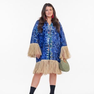 May include: A blue tie-dye dress with long sleeves and fringe trim. The dress has a V-neck and is paired with black boots and a green woven handbag. The model is wearing red earrings.