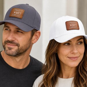 May include: A dark gray baseball cap and a white baseball cap, each featuring a rectangular brown leather patch on the front. The patches are embossed with the text PRAY WAIT TRUST and a small cross symbol centered on the right side of the text.