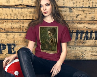 Puede incluir: Camiseta burdeos con una ilustración enmarcada de un esqueleto cuidando plantas, con el texto "PLANT LOVER" en la parte inferior. La camiseta la lleva una persona sentada frente a un fondo de madera.