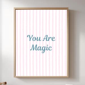 May include: A rectangular wall art print in a light wooden frame featuring a vertical pattern of soft pink and white stripes. Centered on the print is the text "You Are Magic" written in a teal blue script font against the striped background.