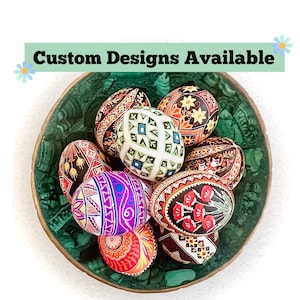May include: A collection of intricately decorated Easter eggs in a green bowl. The eggs feature various patterns and colors, including red, yellow, blue, and purple. The text "Custom Designs Available" is displayed above the eggs.