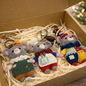 May include: A gift box filled with three handmade crochet mouse keychains. Each mouse is gray with pink ears and various outfits, including a chef's uniform and a patchwork design. The box is lined with straw.