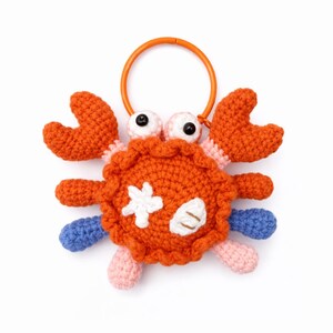 May include: A handmade crochet crab toy in orange, pink, and blue. The crab has large orange claws, googly eyes, and a white starfish and shell design on its body. An orange ring is attached to the top.