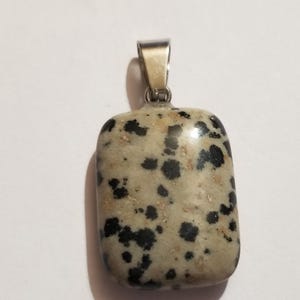 May include: A rectangular pendant made of polished dalmatian jasper stone featuring a speckled pattern of black spots on a beige background, attached to a silver-toned metal bail at the top for hanging on a necklace chain or cord.
