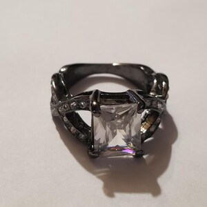 May include: A dark gunmetal-toned ring featuring a large, rectangular, clear faceted center stone held by four prongs. The band is designed with an open, crisscross pattern along the sides, which is accented with small, clear, round stones.