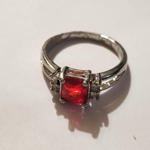 May include: A silver-toned ring featuring a rectangular, faceted red center stone held by four prongs. The band splits into two sections on each side, with three small, clear, round stones set into the metal on both sides of the center stone.