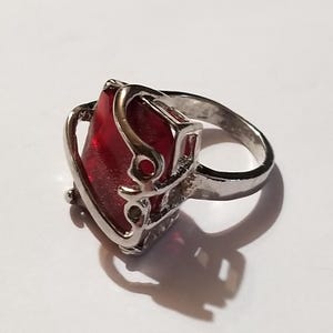 May include: A silver-toned ring featuring a large, rectangular, deep red faceted stone set within a decorative metal frame that curves around the sides of the gem. The band is smooth and metallic, contrasting with the vibrant red center piece.
