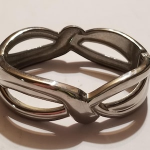 May include: A polished silver-tone metal hinged bangle bracelet featuring an openwork design with interlocking loops that create a continuous infinity-like pattern. The bracelet has a smooth, reflective surface and a secure side hinge mechanism.