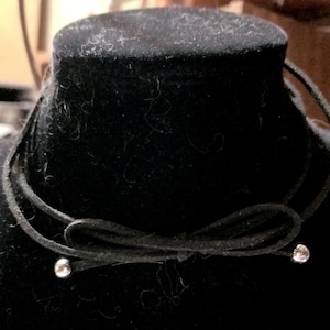 May include: A black suede cord choker necklace featuring a central bow design. The two ends of the cord are finished with small, polished silver-tone metal beads. The necklace is displayed on a black velvet-textured form to show the structure.