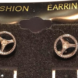 May include: A pair of silver-toned stud earrings shaped like miniature steering wheels, each featuring a central clear rhinestone. The earrings are attached to a black display card with the text FASHION EARRINGS printed in gold lettering.