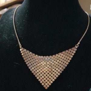May include: A rose gold-toned necklace featuring a triangular pendant composed of small, interlocking circular metal discs. The pendant is attached to a thin, flexible chain that connects to the top corners of the triangular mesh structure.