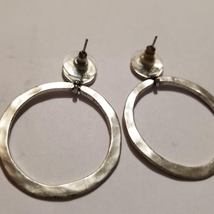 May include: A pair of silver-toned hoop earrings featuring a hammered, irregular circular design. Each earring is attached to a post-style backing with a clear, circular plastic clutch, showcasing a textured metallic finish on the hoop surface.