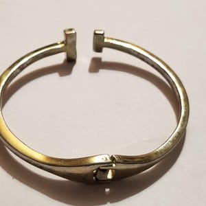 May include: A gold-toned metal cuff bracelet featuring a T-shaped design at the open ends. The bracelet has a hinged mechanism at the bottom, allowing it to open and close securely. The surface has a polished finish with subtle metallic reflections.