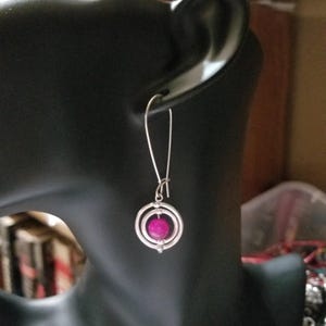 May include: A silver-toned earring featuring a long, slender hook wire attached to a circular pendant. The pendant consists of two concentric silver rings surrounding a single, round, vibrant magenta bead suspended in the center of the circles.