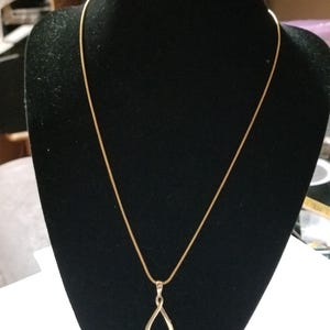 May include: A gold-toned necklace featuring a thin, smooth chain and a teardrop-shaped pendant. The pendant has an open center and a polished finish, suspended from a simple bail. The necklace is displayed on a black stand to highlight the metal.