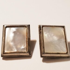 May include: A pair of square cufflinks with a silver-colored frame and a mother-of-pearl center. The cufflinks are set against a white background, showcasing their reflective, iridescent surface.