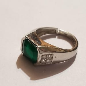 May include: A silver-toned ring featuring a large, rectangular, emerald-green stone set in the center. The band includes a small square accent panel on the side, which is embedded with four tiny, clear, sparkling stones. The ring has an open back.