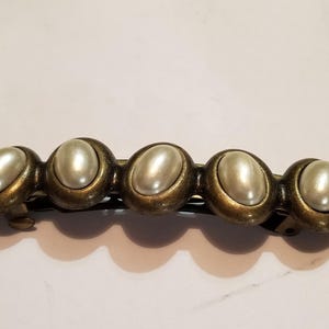 May include: A hair barrette featuring five oval-shaped, cream-colored faux pearls set in a row. Each pearl is encased in a circular, antiqued brass-toned metal frame. The barrette has a hinged metal clasp mechanism on the back for securing hair.