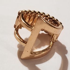 May include: A gold-toned ring featuring a geometric, angular design with a pointed front element. The band is constructed from a series of small, connected metallic spheres that allow for flexibility, creating an adjustable fit for the wearer.
