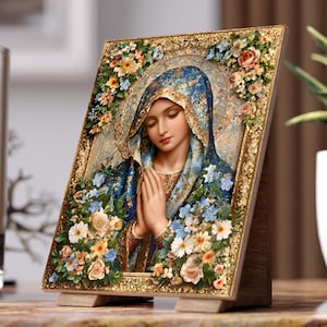 May include: A rectangular plaque featuring a portrait of a figure with closed eyes and hands pressed together in prayer. The figure wears a blue head covering and is surrounded by a decorative gold frame adorned with colorful floral arrangements.