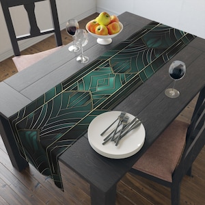 May include: A long table runner featuring a dark green and black geometric pattern with thin gold lines. The runner is placed on a dark wooden table with a stack of white plates, two wine glasses, and a white bowl filled with red and yellow apples.