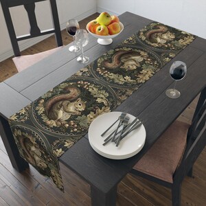 Puede incluir: Un camino de mesa oscuro con un patrón repetitivo de ardillas rodeadas de hojas de roble y bellotas en tonos marrón, verde y dorado. El camino está colocado sobre una mesa de madera oscura con platos blancos, cubiertos y copas de vino.
