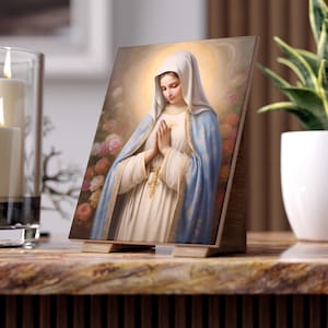 Puede incluir: Impresión enmarcada de una figura religiosa, una mujer con una túnica blanca y azul con las manos juntas. La obra de arte está ambientada sobre un fondo de suaves rosas rosas y un halo dorado. El marco es de madera.