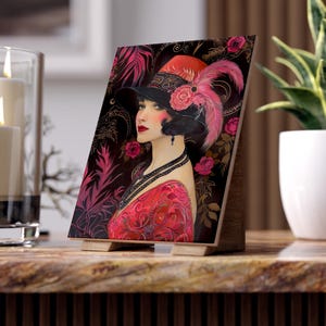 May include: A rectangular art print on a wooden stand featuring a portrait of a person wearing a black hat with a pink feather and a red rose. The person has dark hair, red lipstick, and a black beaded necklace, set against a dark floral background.