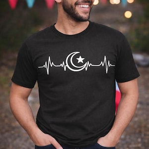 Puede incluir: Camiseta negra con un gráfico blanco de una línea de latido cardíaco con un diseño de luna creciente y estrella. La camiseta es de cuello redondo y manga corta.