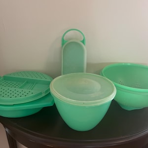 May include: A collection of vintage mint green kitchenware, including a colander, a bowl, a container with a lid, and a rectangular container with a handle. The items are made of plastic and are arranged on a dark surface.