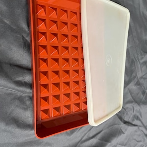 May include: A rectangular, red plastic tray with a textured, geometric pattern on the inside. A white, rectangular lid sits partially on the tray. The tray and lid are on a gray surface.