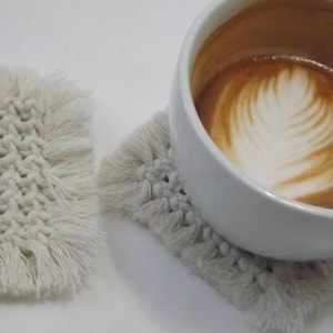 May include: A white ceramic coffee cup with latte art sits on a square, off-white macrame coaster. Another macrame coaster is next to the cup. The coasters have a woven texture with fringe.