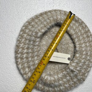 May include: A beige and white speckled beret with a tape measure showing a diameter of approximately 11 inches. The beret has a soft, fuzzy texture and a tag that reads "ANTHROPOLOGIE".