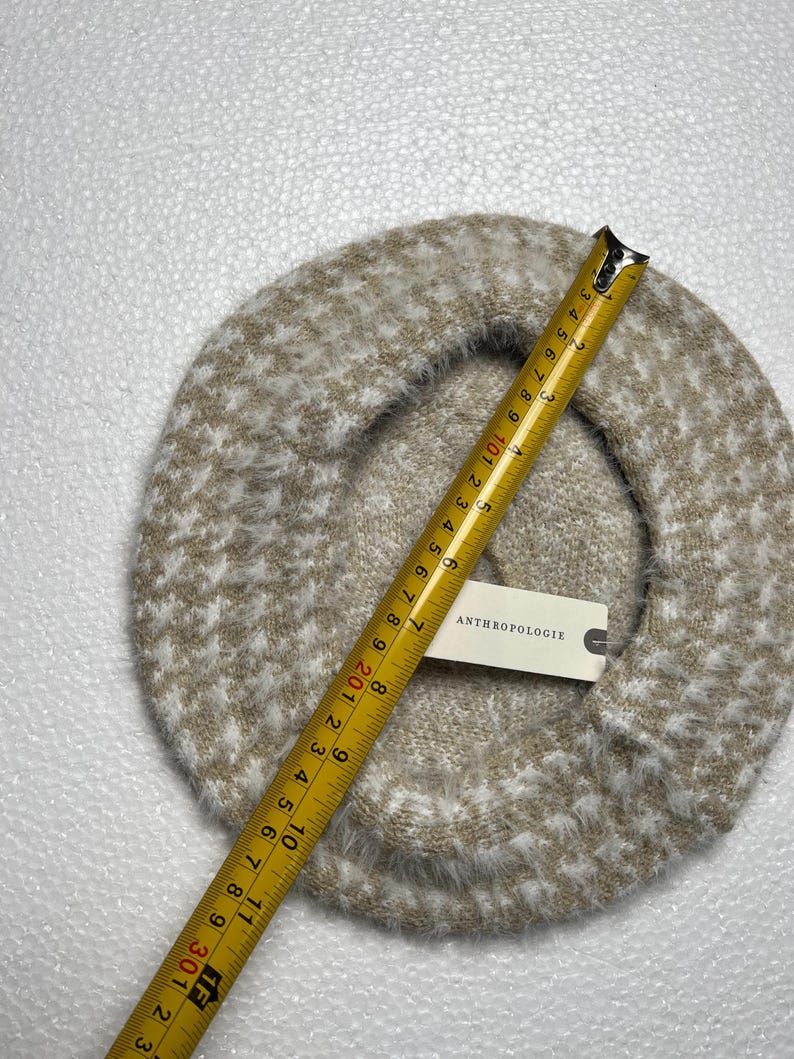 May include: A beige and white speckled beret with a tape measure showing a diameter of approximately 11 inches. The beret has a soft, fuzzy texture and a tag that reads "ANTHROPOLOGIE".