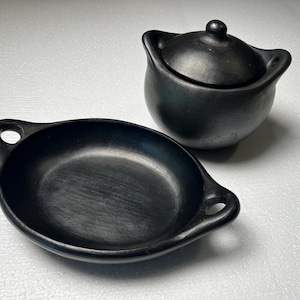 May include: Two pieces of black pottery. One is a small, round pot with a lid and two handles. The other is a shallow, oval dish with two handles. Both pieces have a matte finish and appear handmade.