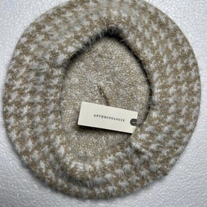 May include: A beige and white patterned beret with a soft, fuzzy texture. The beret has a houndstooth-like design and a tag that reads "ANTHROPOLOGIE". This stylish hat is perfect for adding a touch of sophistication to any outfit.