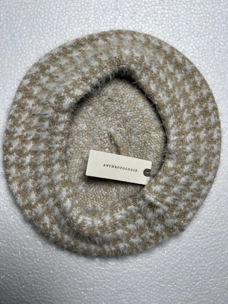 May include: A beige and white patterned beret with a soft, fuzzy texture. The beret has a houndstooth-like design and a tag that reads "ANTHROPOLOGIE". This stylish hat is perfect for adding a touch of sophistication to any outfit.