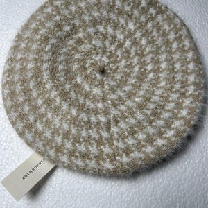 May include: A beige and white houndstooth pattern beret. The beret is round with a soft, fuzzy texture and a small tag that reads "ANTHROPOLOGIE".