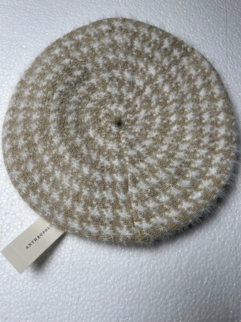 May include: A beige and white houndstooth pattern beret. The beret is round with a soft, fuzzy texture and a small tag that reads "ANTHROPOLOGIE".