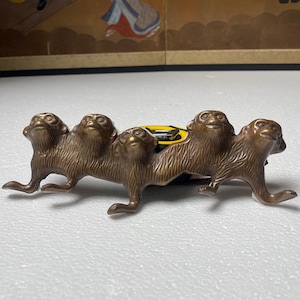 Puede incluir: Un perchero de pared de metal color bronce con cuatro figuras de monos esculpidas conectadas en fila. Los monos muestran texturas de pelaje detalladas y expresiones mirando hacia arriba. Se observa una cinta métrica amarilla detrás.