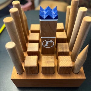 May include: A wooden game with a brown base, small wooden cubes, and tall wooden dowels. A central wooden piece has a blue crown and a white "F" logo. The game is designed for strategic play and skill development.