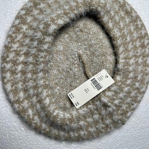May include: A beige and white houndstooth pattern beret. The hat is made of a soft, fuzzy material. A white tag is attached to the beret.