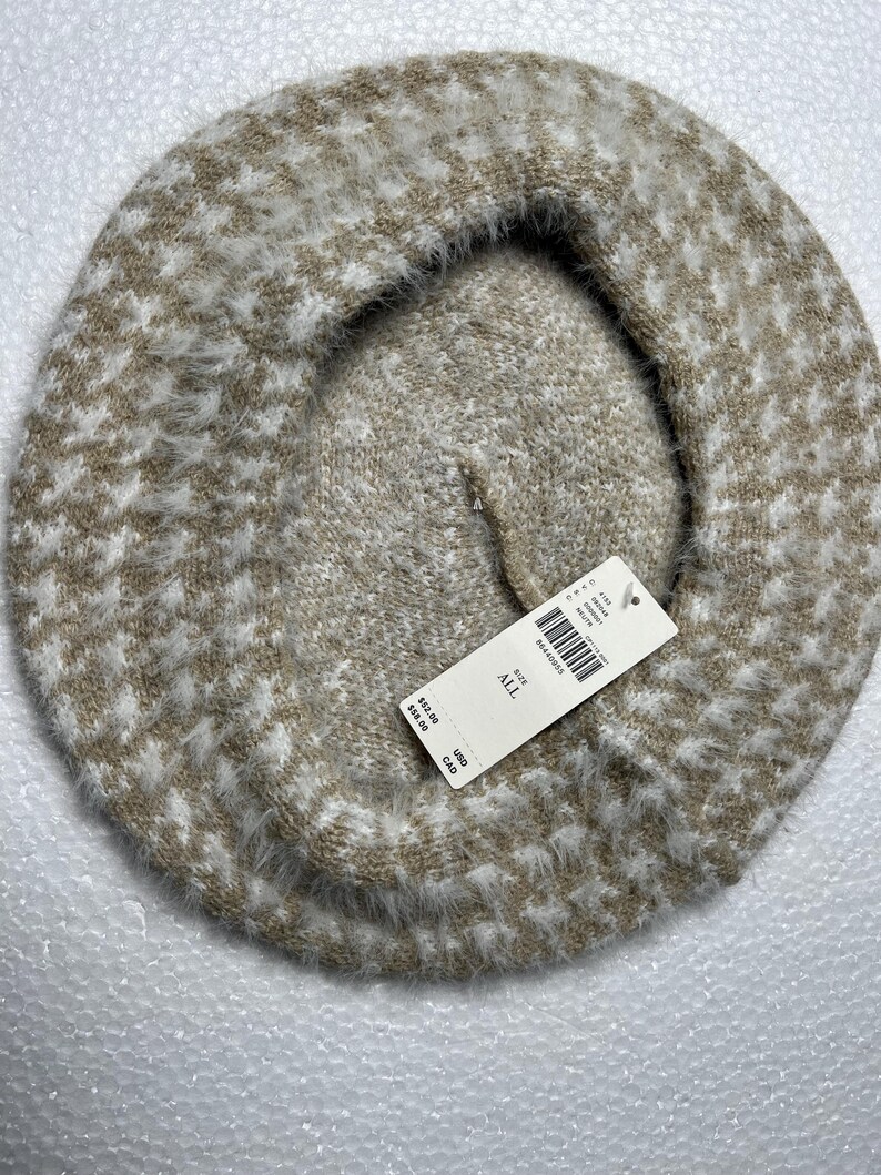 May include: A beige and white houndstooth pattern beret. The hat is made of a soft, fuzzy material. A white tag is attached to the beret.