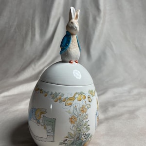 May include: A white ceramic egg-shaped jar with a lid topped by a sculpted Peter Rabbit figure wearing a blue jacket. The jar features a colorful wrap-around illustration of Peter Rabbit in bed, garden flowers, pears, and a watering can.