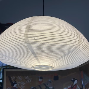 May include: A white paper pendant lamp with a rounded, flattened shape and horizontal ribbed texture. The light fixture features a thin wire frame visible at the bottom opening and is suspended by a single black cord from the ceiling above.