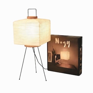 May include: A square paper lantern table lamp with a ribbed texture, a wooden base, and three thin black metal legs. A black metal handle sits on top. Next to the lamp is a black cardboard box featuring the text Nogy and an image of the lamp.