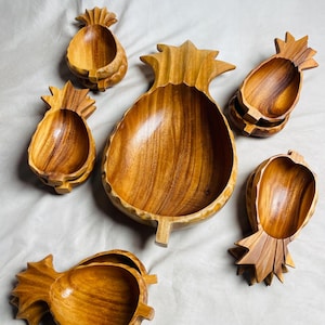 May include: A collection of six pineapple-shaped wooden bowls of varying sizes. Each bowl features a smooth, polished finish with visible wood grain patterns, carved into the distinct form of a pineapple with a leafy top and a rounded base.
