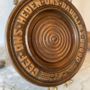 May include: A round, wooden decorative plate with carved concentric circles and the words "GEEF ONS HEDEN ONS DAGELIJKSE BROOD" in raised lettering. The plate is brown with gold-colored text and a wheat stalk design. It is displayed on a brass stand.