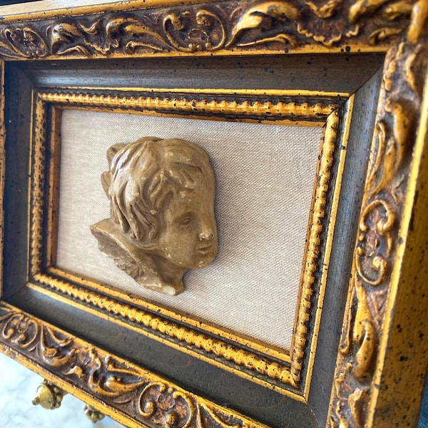 Puede incluir: Una decoración de pared enmarcada con un marco dorado adornado y un fondo beige. El marco tiene un borde interior marrón oscuro. Un relieve de yeso de la cabeza de un querubín está centrado en el marco.